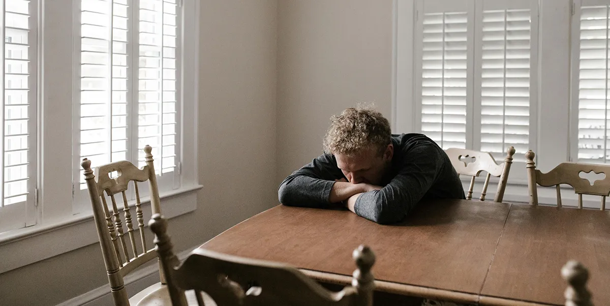 A man sits alone at a dining table, head resting on his arms, highlighting the struggles of loneliness and the importance of addressing mental health issues