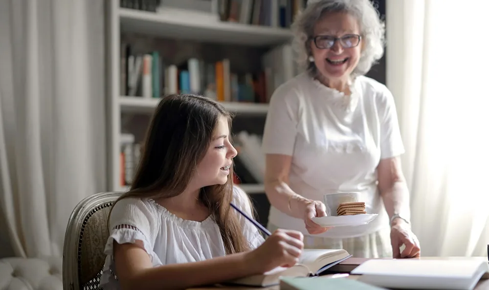 An elderly woman smiling as she serves a cup of tea and biscuits to a young girl who is in an online therapy session.