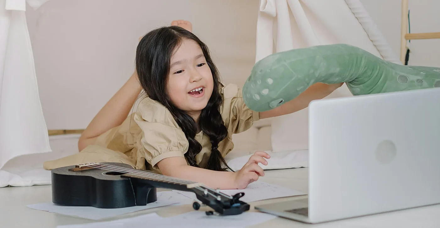 A child engaged in therapy play, lying on the floor with a stuffed toy, a ukulele, and sheets of paper, smiling while interacting with a laptop