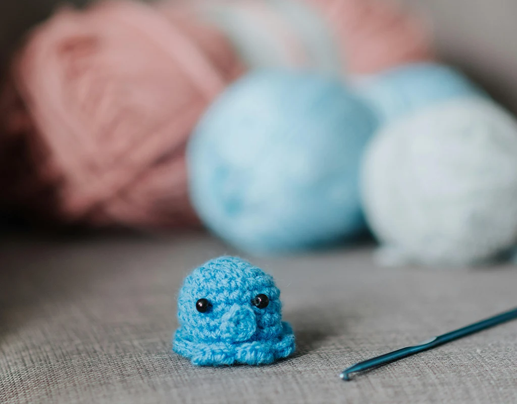 A small light blue crocheted octopus on a sofa with blurred cotton yarn in the background