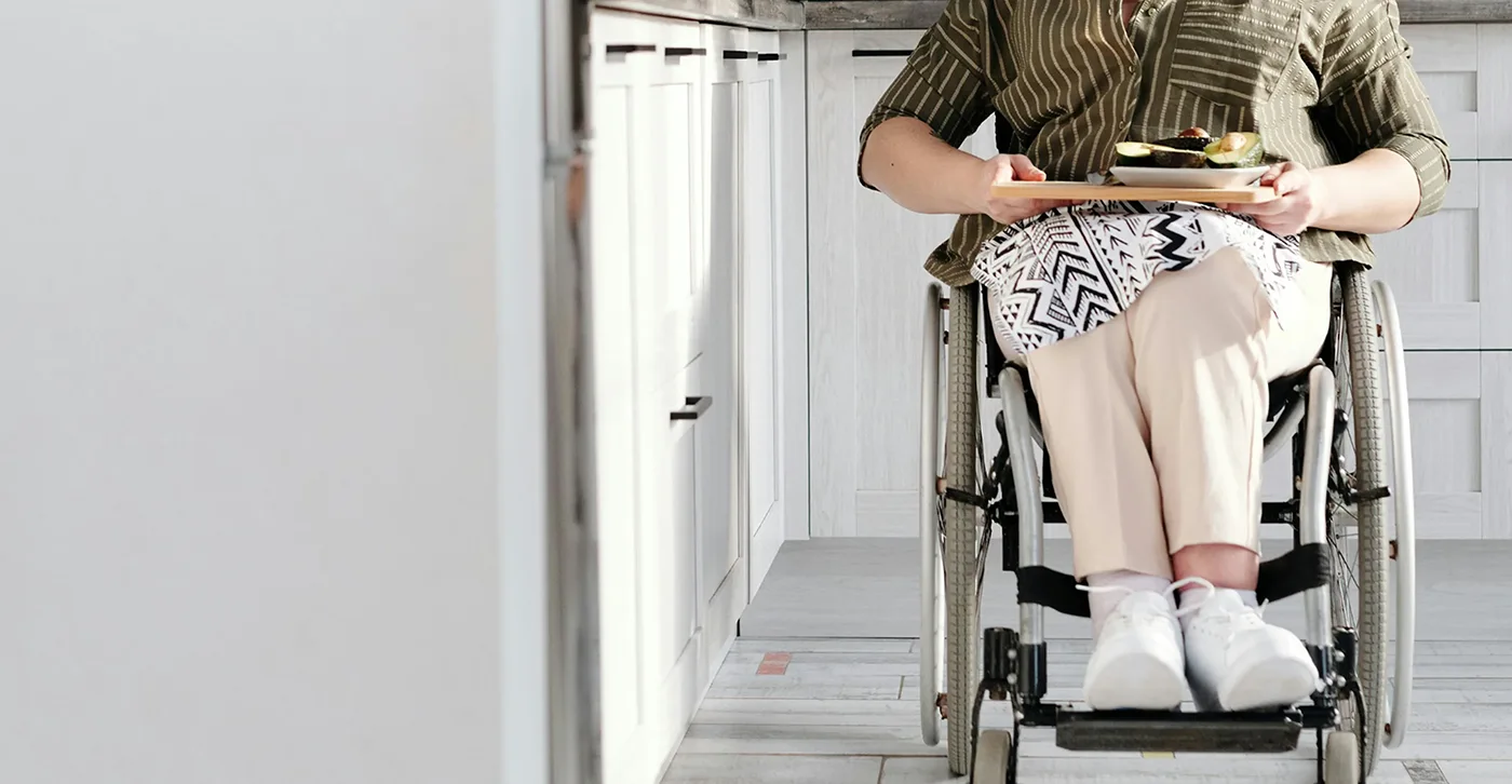 person in a wheelchair balancing a tray with food, moving through a kitchen with light-colored cabinets and flooring