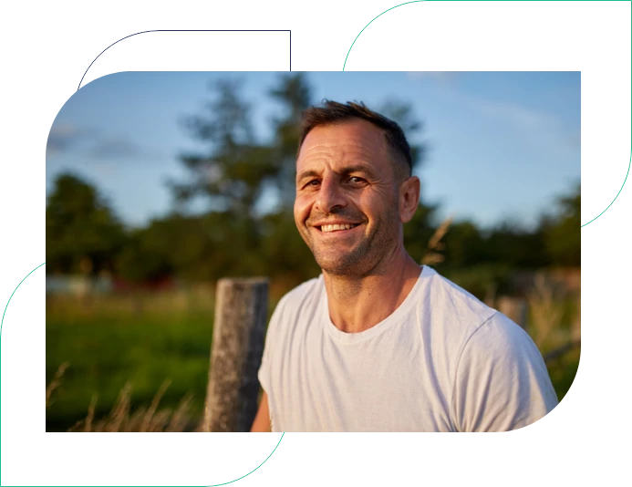 A man wearing a white t-shirt stands confidently in a green field surrounded by nature