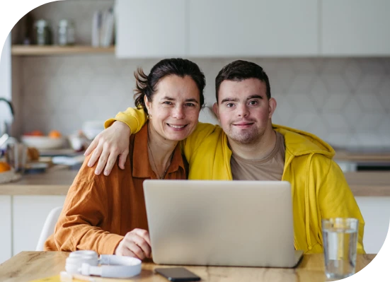 A man with Down syndrome and a woman sit together at a table, with a laptop in front of them.