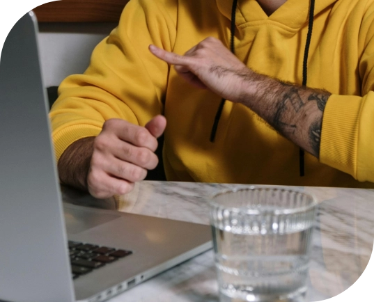 A person in a yellow hoodie using sign language in his NDIS counselling session while sitting at a marble table with a laptop.