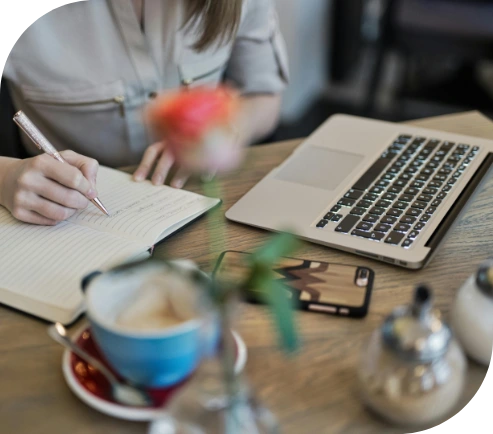 A woman writes in a notebook next to her laptop while having coffee, receiving occupational therapy for mental health to improve her daily functioning and well-being.