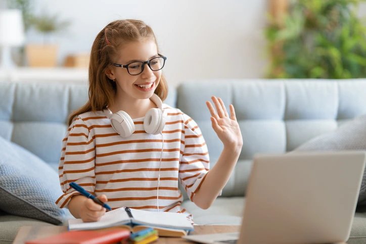 A young girl waves during an online telehealth occupational therapy session, wearing headphones and taking notes from home.