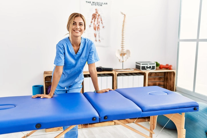 Smiling physiotherapist standing by a treatment table, ready for a therapy session to support patients in physical rehabilitation.