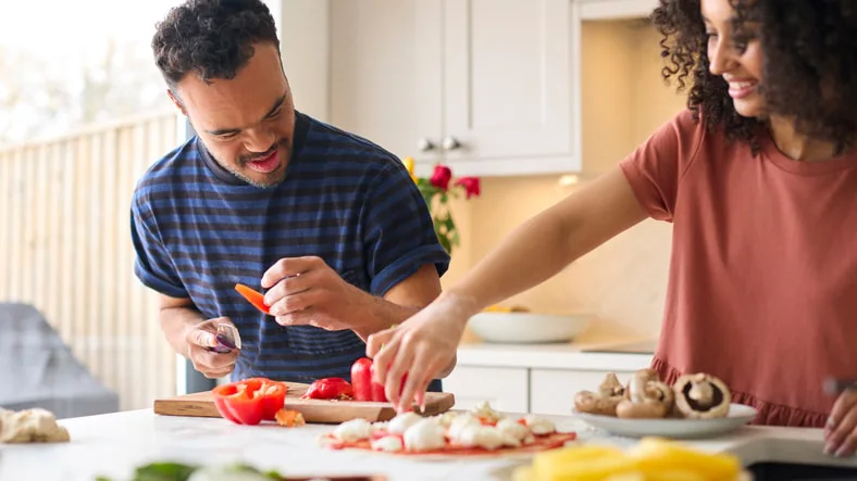 Two people in a kitchen are preparing vegetables. One is slicing a red pepper, while the other arranges ingredients on a cutting board.